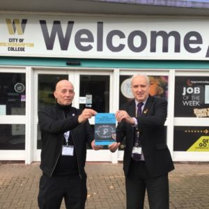 Author Paul Cunningham, left, wearing a black sweater and trousers, and Ian Browne, vice principal wearing a dark suit with blue shirt and dark tie, are holding a copy of Paul's book with a blue cover