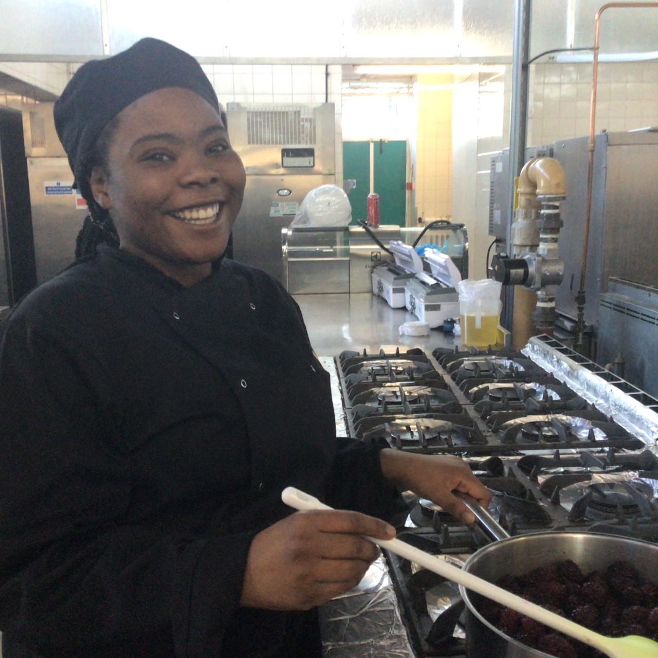 Cookery student Michelle Murderere wearing a dark uniform, holding a wooden spoon and stirring contents of a saucepan