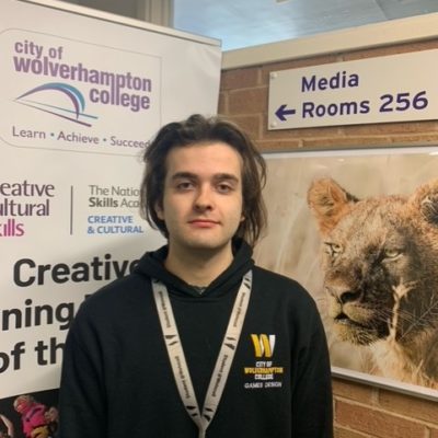 Games design student Adam Cater, wearing a black hoody with the college logo in mustard and yellow on the left, standing in front of a creative arts stand