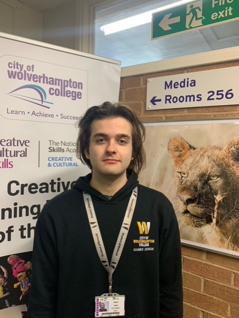 Games design student Adam Cater, wearing a black hoody with the college logo in mustard and yellow on the left, standing in front of a creative arts stand