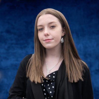 Photograph of performing arts student kacey Howell, wearing a black and white spotted top, standing in front of a blue backdrop