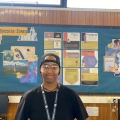 Student Dalila Madiera wearing a black cap and black top, standing in front of a notice board