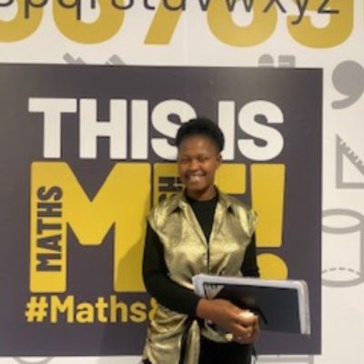 Business admin student Edette Esoh wearing a gold jacket, standing in front of a black and yellow college sign