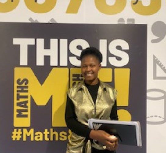 Business admin student Edette Esoh wearing a gold jacket, standing in front of a black and yellow college sign