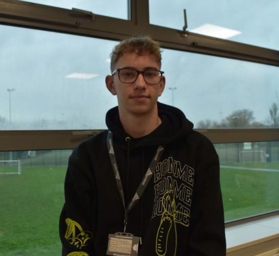 Sport student Lewis Smith wearing a black hoodie and a college lanyard, standing in front of a window overlooking the sports pitch