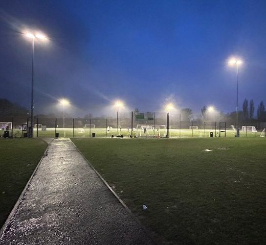 Picture looking across the football pitch at six illuminated floodlights against a dark sky