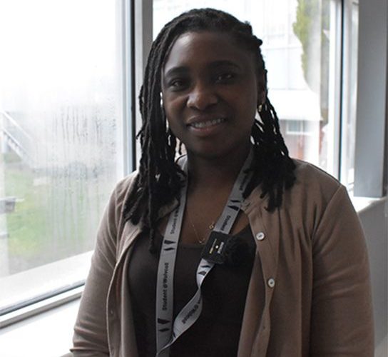 Tapiwa Hakutangwi, wearing a black shirt and brown jacket standing in front of a window