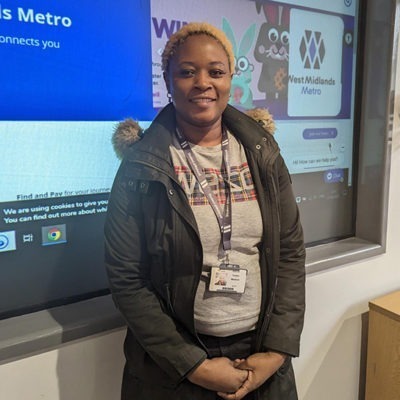 Yeukai Makinzi wearing a grey top and black coat, standing in front of a display board giving details of careers with Midland metro