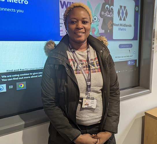 Yeukai Makinzi wearing a grey top and black coat, standing in front of a display board giving details of careers with Midland metro