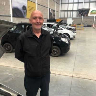 Former student Adrian Howard wearing dark blue overalls standing in front of vehicles in the workshop