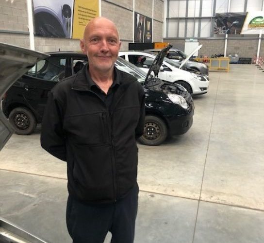 Former student Adrian Howard wearing dark blue overalls standing in front of vehicles in the workshop