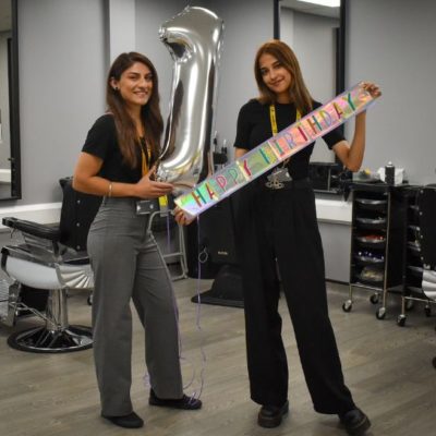 Two members of staff in the college hair salon - one holding a large silver '1' and th other holding a colourful happy birthday banner