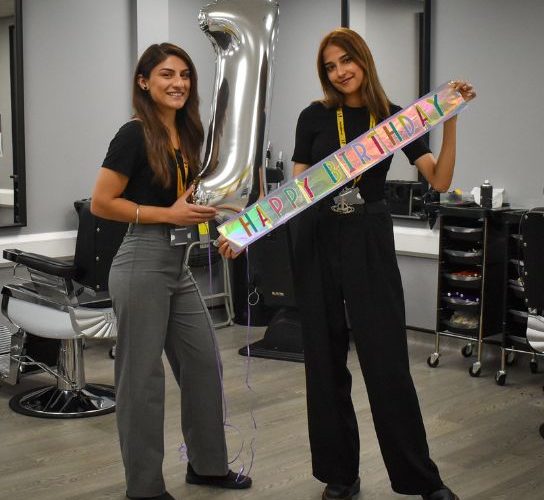 Two members of staff in the college hair salon - one holding a large silver '1' and th other holding a colourful happy birthday banner