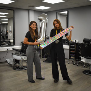 Two members of staff in the college hair salon - one holding a large silver '1' and th other holding a colourful happy birthday banner