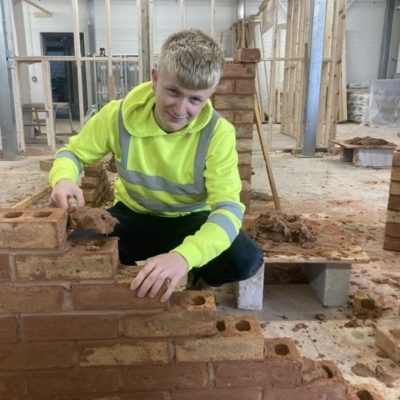 Bricklaying apprentice Charlie Cole, wearing a fluorescent jacket building a wall in the bricklaying workshop