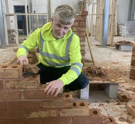 Bricklaying apprentice Charlie Cole, wearing a fluorescent jacket building a wall in the bricklaying workshop