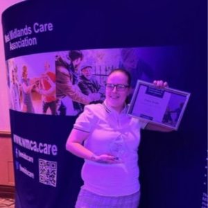 Claire Smith wearing a white top holding an award and certificate in front of a West Midlands Care Association stand.