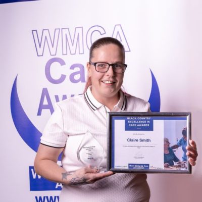 Award winner Claire Smith wearing a white top, standing in front of a West Midalnds Care Association banner, holding a certificare and glass trophy