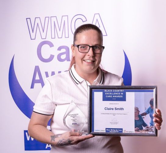 Award winner Claire Smith wearing a white top, standing in front of a West Midalnds Care Association banner, holding a certificare and glass trophy